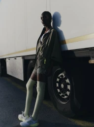A Black woman in an olive jacket, pale green knee-high socks, and sneakers leans against the side of a large white truck in direct sunlight.