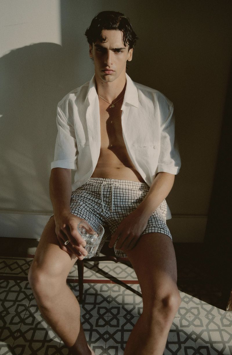Adult male in an unbuttoned white shirt and checkered shorts sits on a chair holding a glass, bathed in warm sunlight.