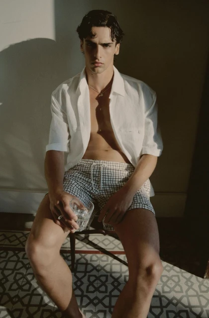Adult male in an unbuttoned white shirt and checkered shorts sits on a chair holding a glass, bathed in warm sunlight.
