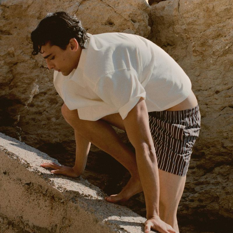 Athletic man climbs a rock face near the ocean. He wears striped shorts and a white shirt in an outdoor setting during the day.