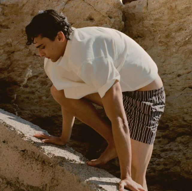 Athletic man climbs a rock face near the ocean. He wears striped shorts and a white shirt in an outdoor setting during the day.