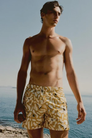 Male fashion model poses in patterned swim trunks against a blue ocean and sky backdrop, showcasing summer beach style.