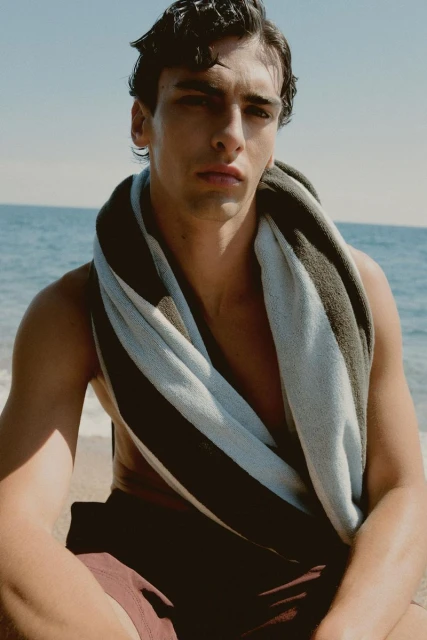 Portrait of a man with dark hair and a towel around his shoulders, sitting on a beach with ocean waves in the background.
