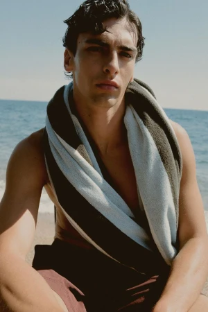 Portrait of a man with dark hair and a towel around his shoulders, sitting on a beach with ocean waves in the background.