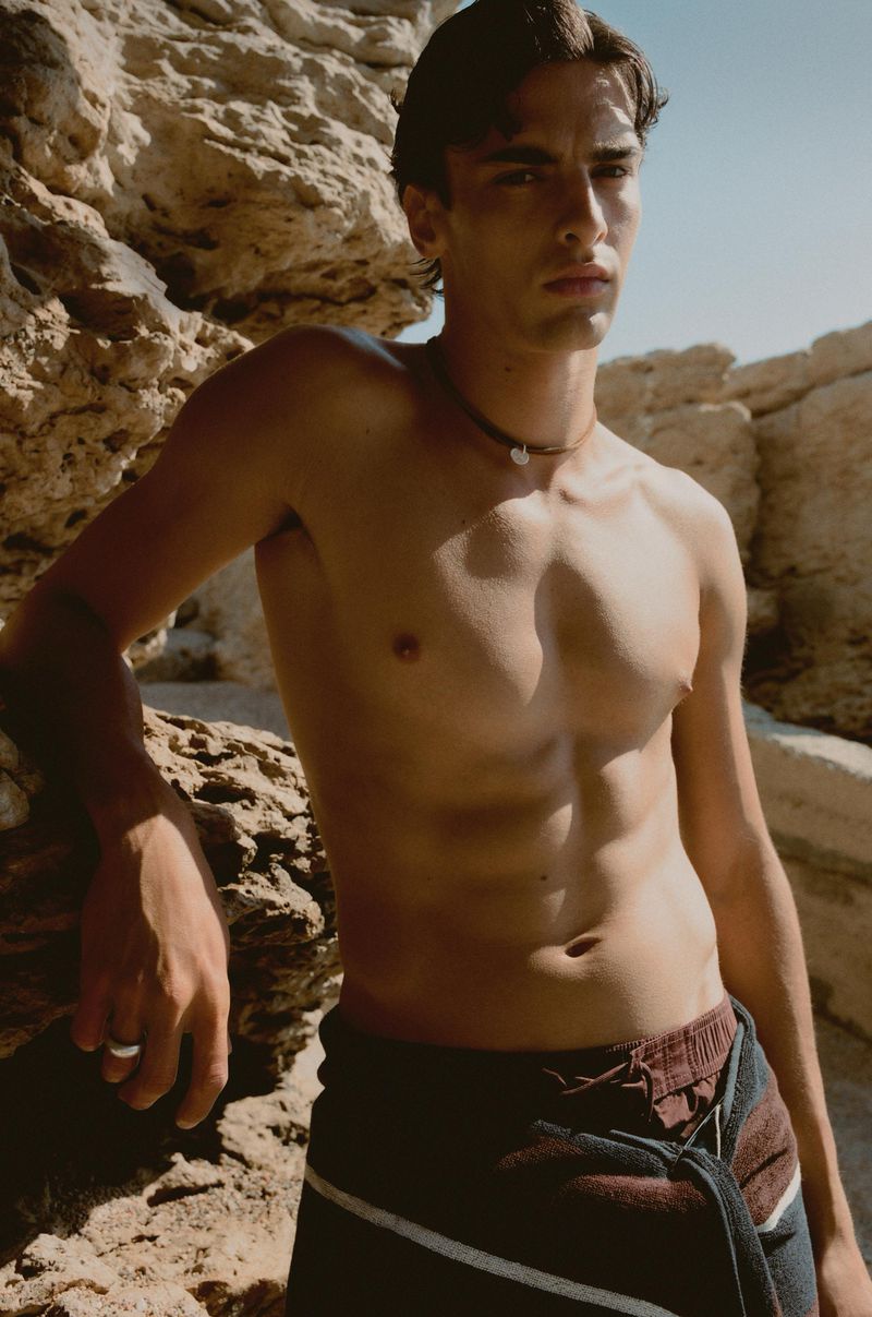 Shirtless male model wearing dark swim shorts and a cord necklace posing outdoors against sunlit rocky cliffs.