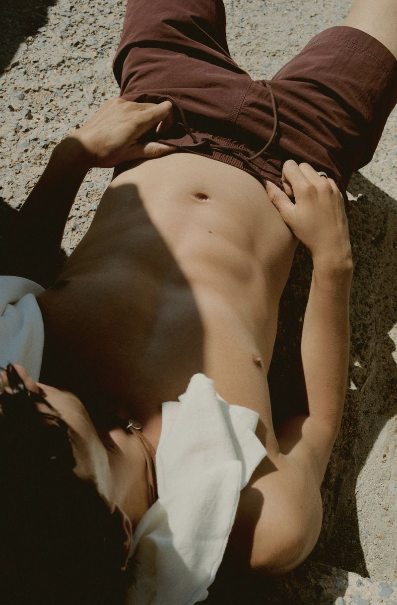 Top-down view of a shirtless person in dark red shorts lying on sunlit concrete with a white cloth, creating a casual, summery vibe.