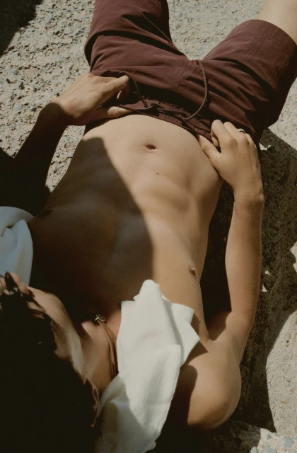 Top-down view of a shirtless person in dark red shorts lying on sunlit concrete with a white cloth, creating a casual, summery vibe.