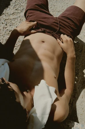 Top-down view of a shirtless person in dark red shorts lying on sunlit concrete with a white cloth, creating a casual, summery vibe.