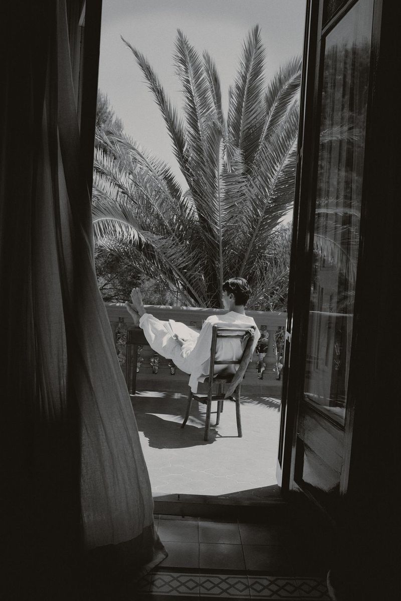 A man in a white robe reclines on a balcony chair with his feet propped up, framed by open doors and palm trees in monochrome.