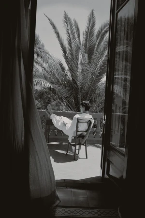 A man in a white robe reclines on a balcony chair with his feet propped up, framed by open doors and palm trees in monochrome.