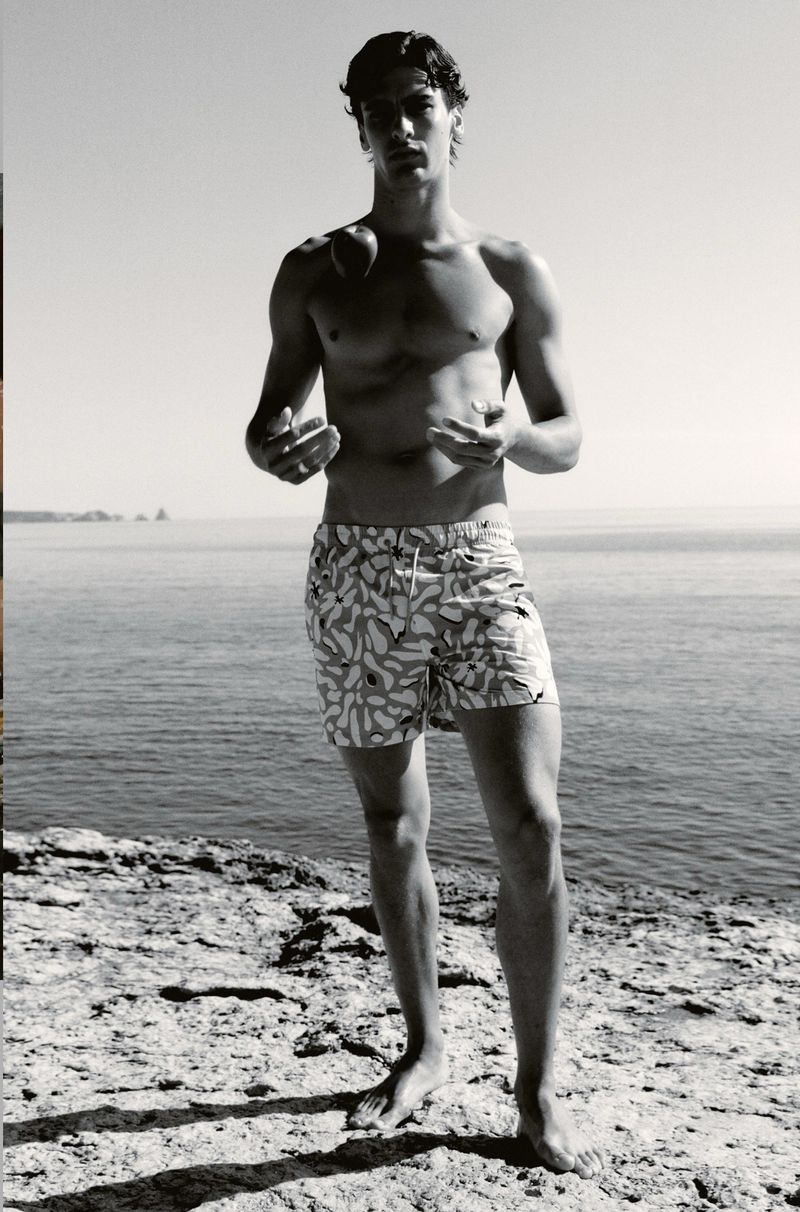 Young man poses in patterned swim trunks by the ocean on a rocky coast in black and white, looking at the camera in an eye-level shot.