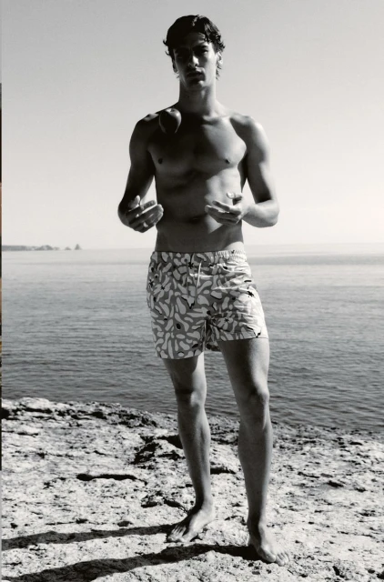 Young man poses in patterned swim trunks by the ocean on a rocky coast in black and white, looking at the camera in an eye-level shot.