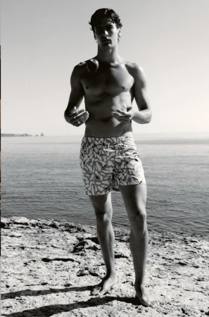 Young man poses in patterned swim trunks by the ocean on a rocky coast in black and white, looking at the camera in an eye-level shot.