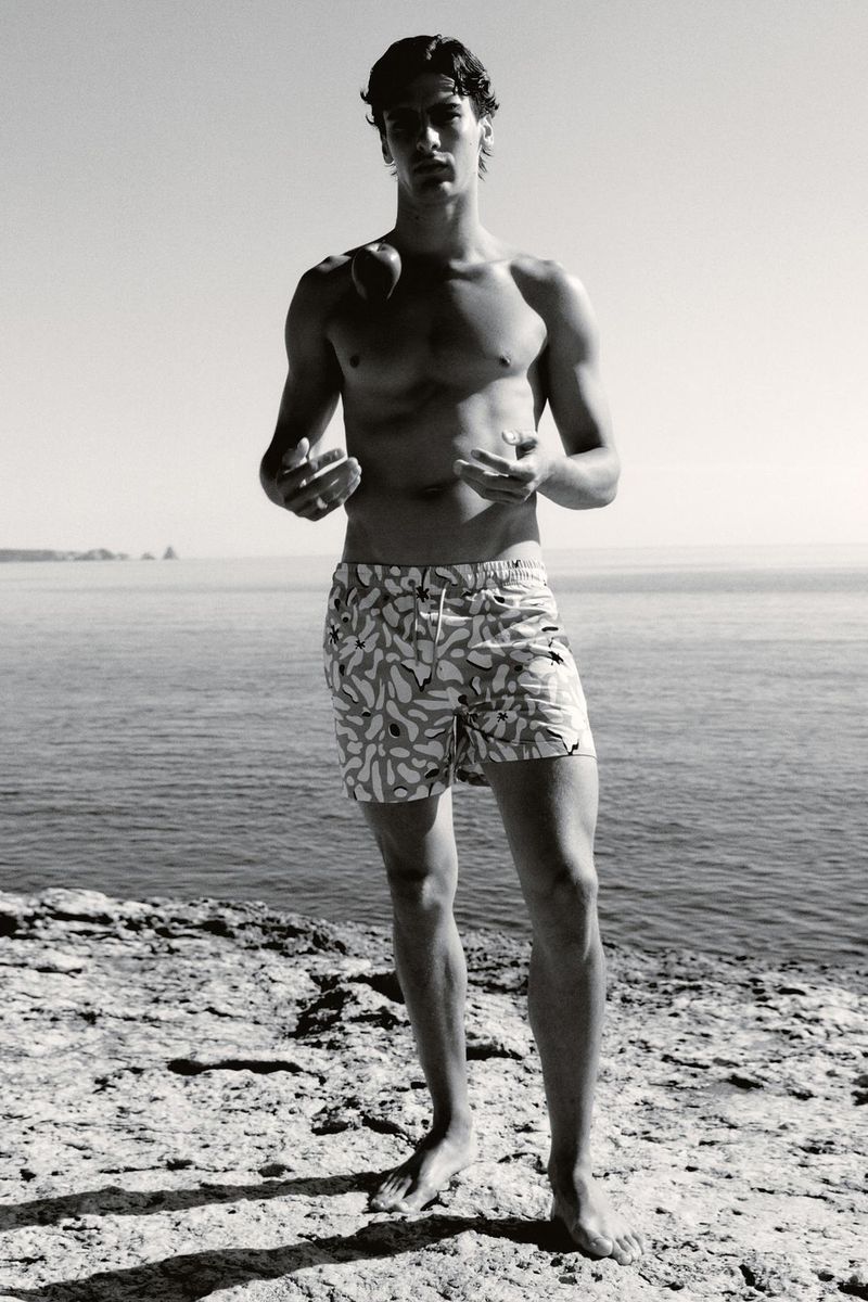 Monochrome photo of a shirtless man in patterned swim trunks standing on rocks by the sea on a sunny day. Fashion or travel concept.