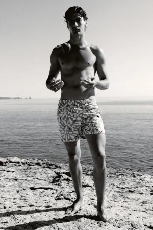 Monochrome photo of a shirtless man in patterned swim trunks standing on rocks by the sea on a sunny day. Fashion or travel concept.