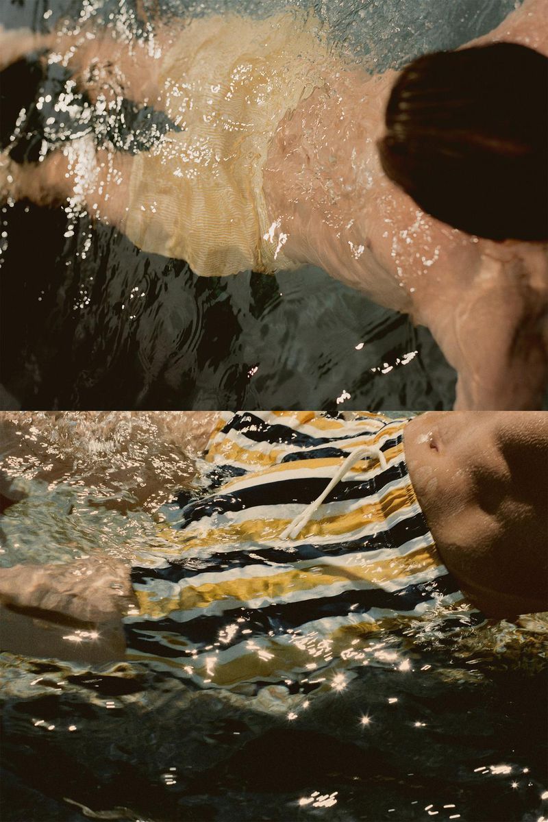 Torso of an adult male swimming in dark water with a striped yellow, white, and black swimsuit visible, partially submerged in the water.