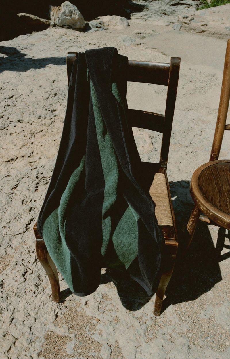 Green and black striped fabric draped on a wooden chair outdoors in natural light, next to a second empty chair on textured ground.