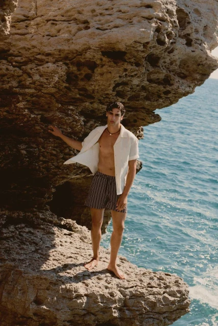 A tanned man stands on rocks in swim trunks and an unbuttoned shirt next to the ocean. He is posing in sunlight.