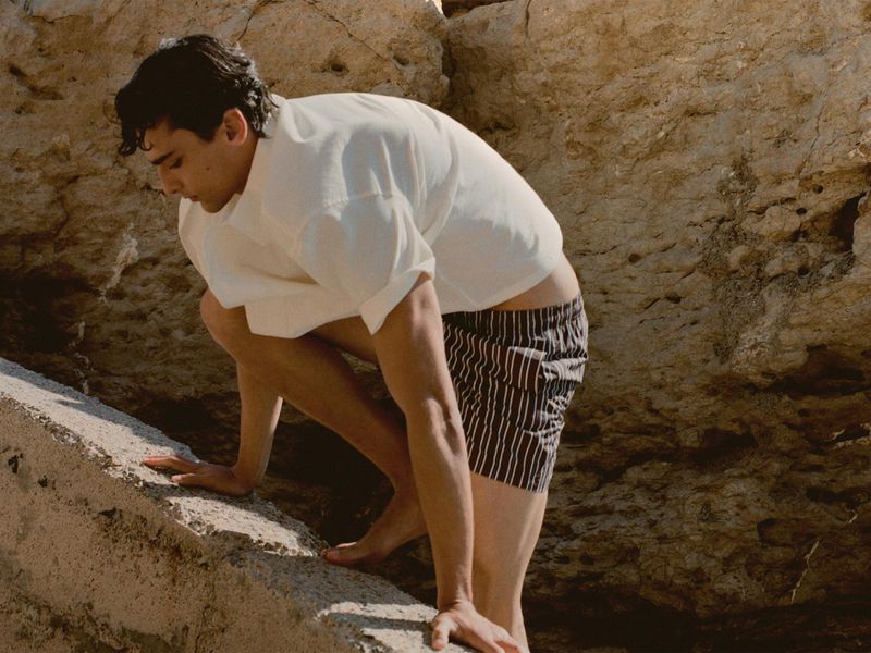 Man in white shirt and striped shorts climbs a light tan rock formation outdoors on a sunny day in a fashion or lifestyle photograph.