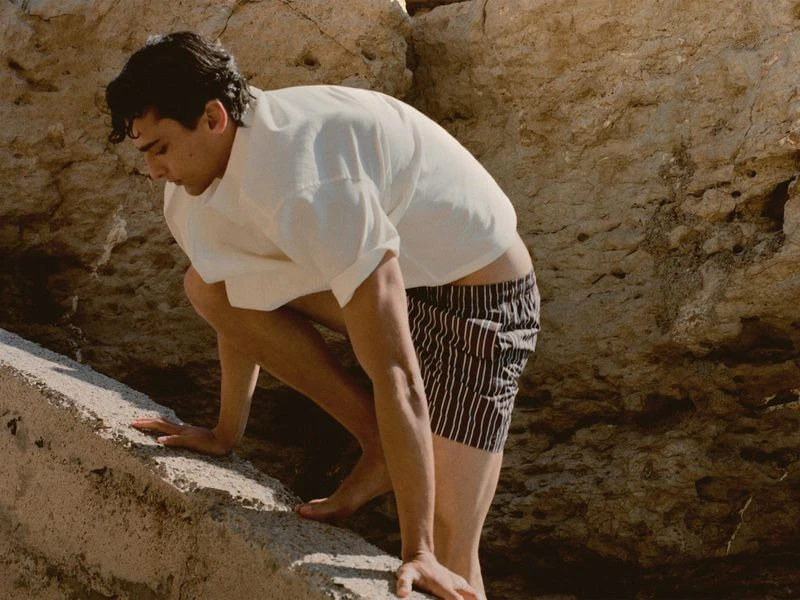 Man in white shirt and striped shorts climbs a light tan rock formation outdoors on a sunny day in a fashion or lifestyle photograph.