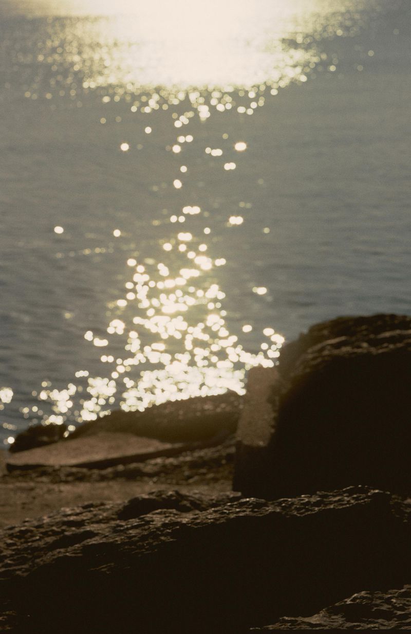 Sunlight sparkles on the ocean surface, reflecting off gentle waves near a rocky shoreline on a bright, sunny day.
