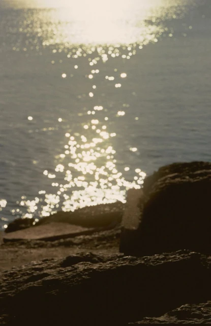 Sunlight sparkles on the ocean surface, reflecting off gentle waves near a rocky shoreline on a bright, sunny day.