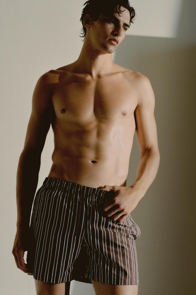 A shirtless adult male model poses in striped shorts, lit by studio lighting, creating a striking portrait in a neutral-toned environment.