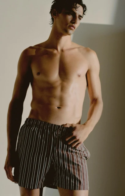 Portrait of a slender adult male model in striped boxer shorts, posing indoors in a neutral studio with soft, diffused lighting.