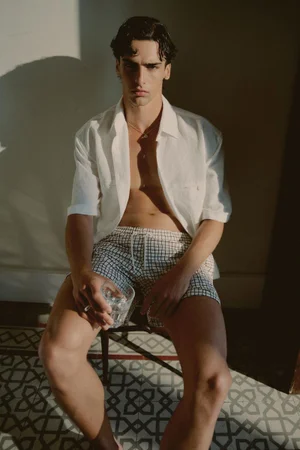 Model Francesco Cicerchia seated in an open white shirt and patterned swim trunks holding a glass of iced water in direct sunlight.