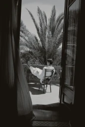 A man in a white robe reclines on a balcony chair with his feet propped up, framed by open doors and palm trees in monochrome.