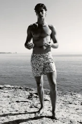 Black and white photo of a shirtless male model in patterned swim trunks standing on a rocky coast overlooking the sea.
