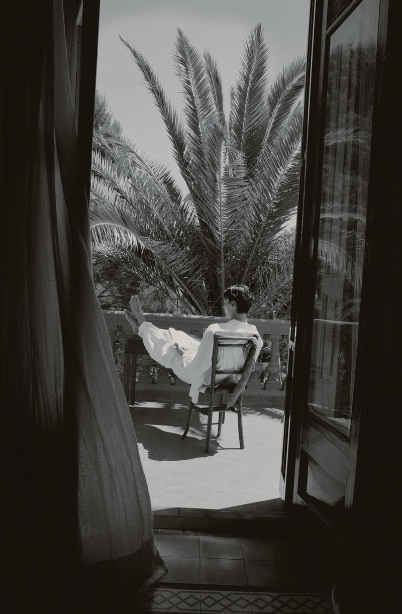 Monochrome image of a person seated on a chair with feet up on a balcony railing framed by open doors and palm trees.
