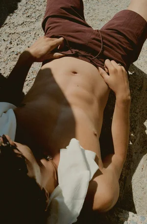 Top-down shot of a shirtless man lying on rocky ground wearing brown drawstring swim shorts with a white towel draped around his neck.