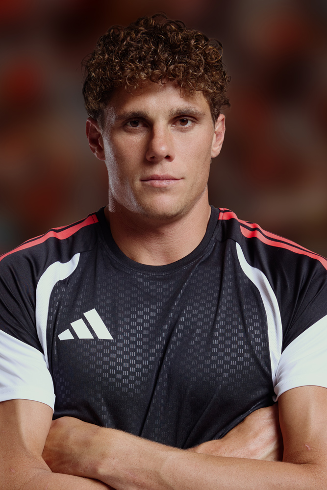 Close-up studio shot of an adult male with curly hair and arms crossed, wearing a sport shirt with an Adidas logo. Neutral expression.