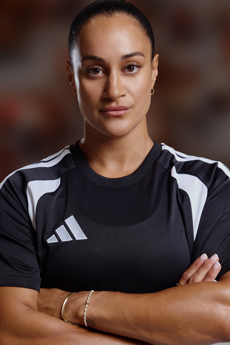Athletic woman with slicked-back hair in a black Adidas short-sleeve jersey with white stripes posing with arms crossed.