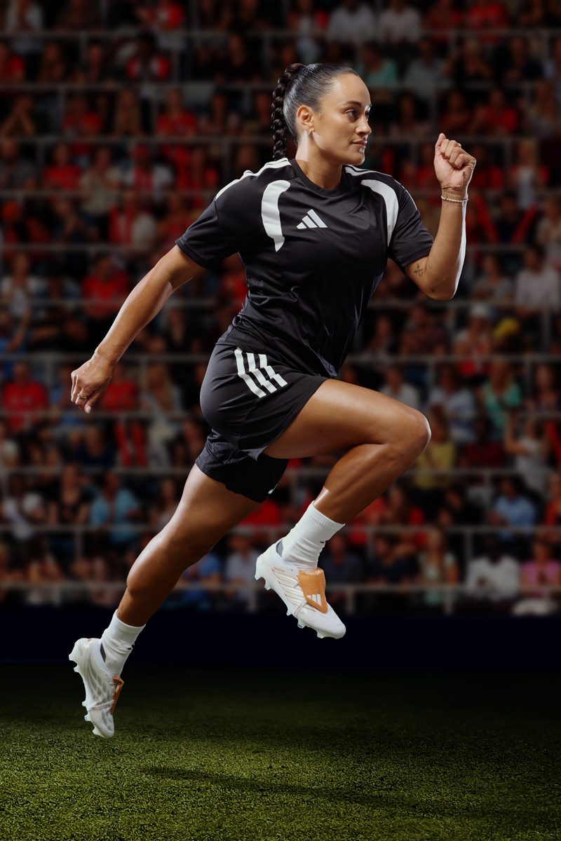 Female soccer player in mid-air sprint wearing a black Adidas kit and white cleats against a stadium crowd background.