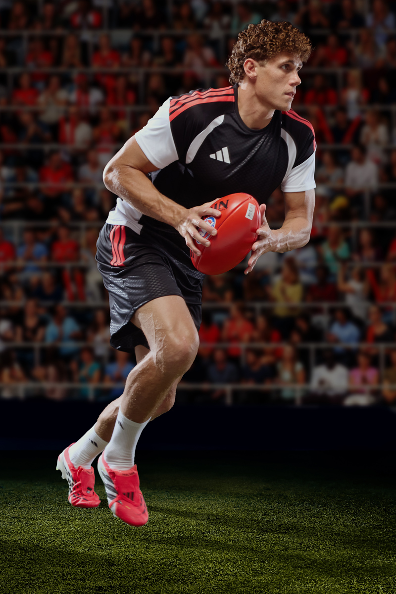 Athlete running on grass holding an Australian rules football in Adidas jersey and bright pink boots against a stadium backdrop.