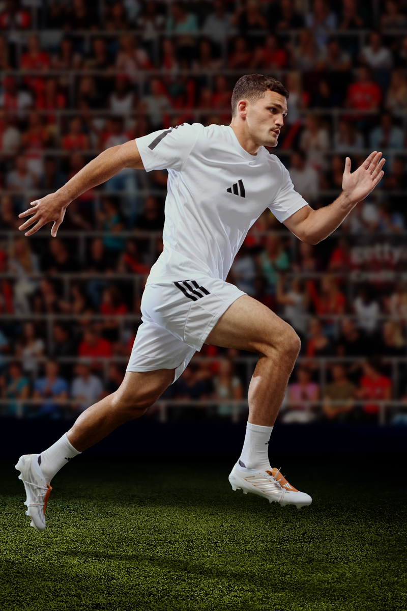 Male athlete in white Adidas sportswear sprinting across a green pitch against a stadium background