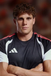 A man with curly brown hair wearing an Adidas black jersey with white and red shoulder stripes, posing with arms crossed.