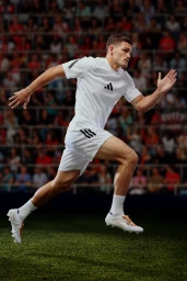 Male athlete in white Adidas sportswear sprinting across a green pitch against a stadium background