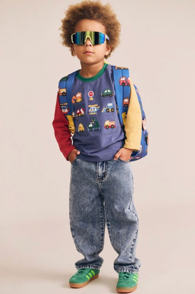 Child wearing shield sunglasses, a vehicle print colorblock shirt, baggy acid-wash jeans, and green sneakers against a beige background.