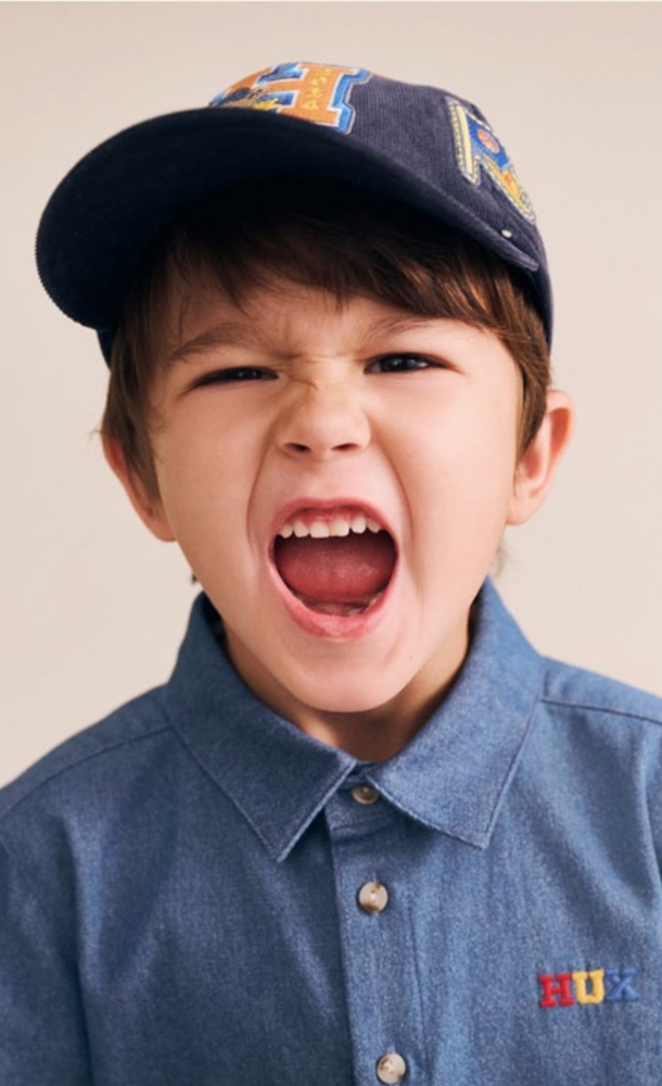 Young boy shouting with mouth open, wearing a blue denim collared shirt and baseball cap against a neutral backdrop.