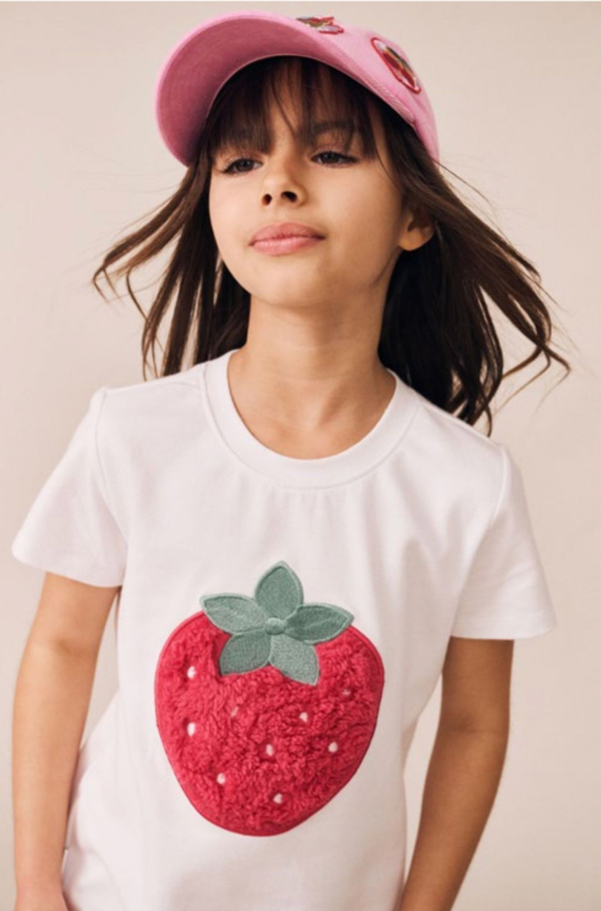 Young girl wearing a pink cap and a white t-shirt featuring a large textured strawberry patch on a neutral studio background.