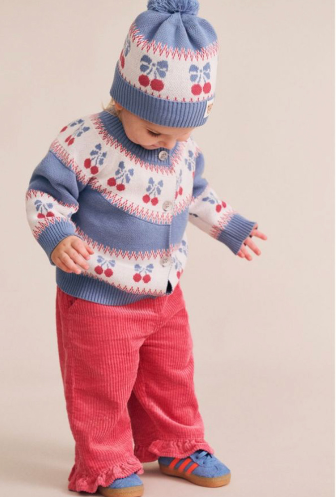 Toddler wearing a blue and white cherry knit cardigan, matching pom-pom beanie, and pink corduroy pants against a studio backdrop.