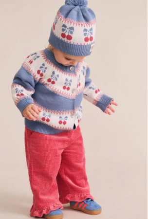 Toddler wearing a blue and white cherry knit cardigan, matching pom-pom beanie, and pink corduroy pants against a studio backdrop.