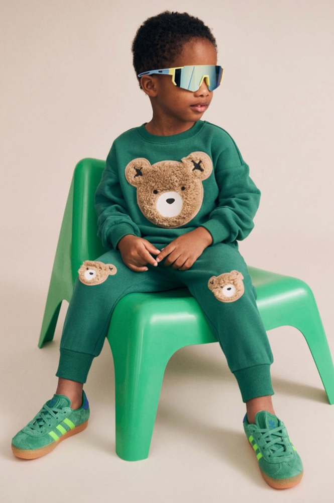 Young boy wearing sporty sunglasses and a green tracksuit with teddy bear patches while sitting on a green plastic chair.