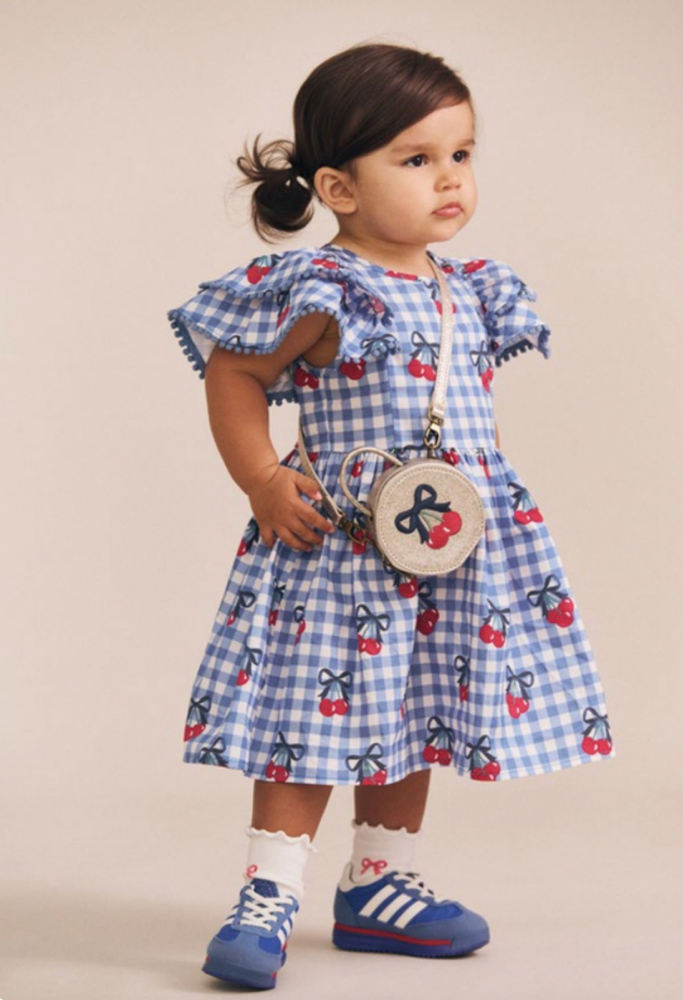 Toddler standing in a blue and white gingham cherry-print dress with a round mini crossbody bag against a neutral studio backdrop.