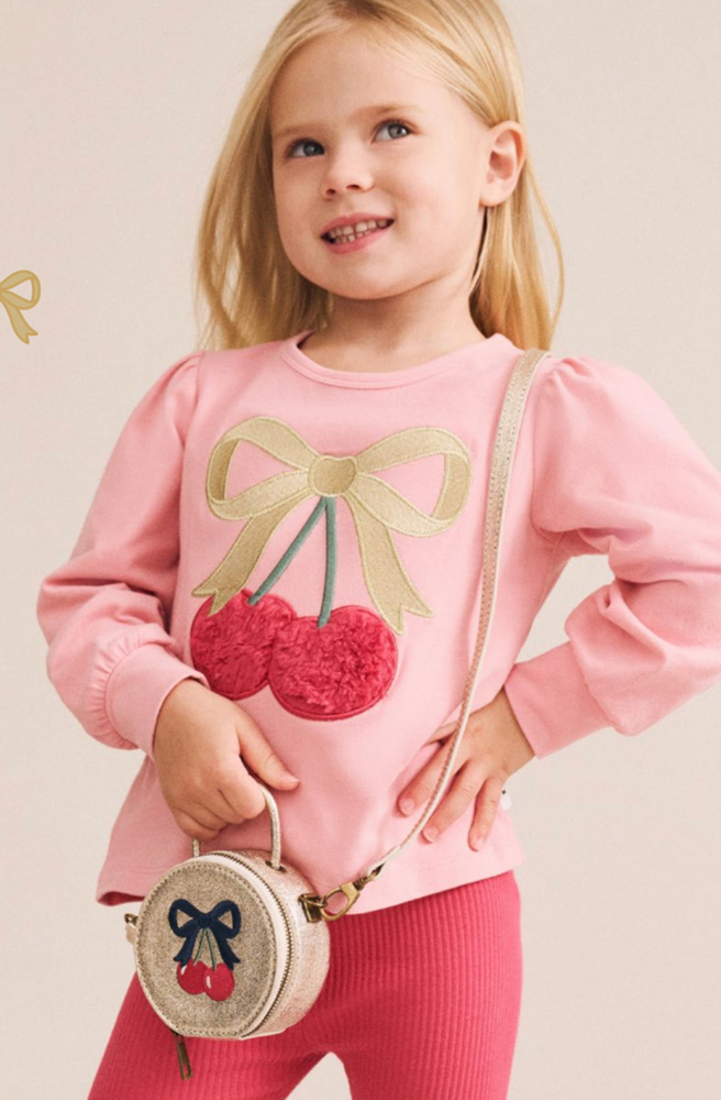 Young child wearing pink long-sleeve top with cherry graphic and pink ribbed pants holding a round metallic bag