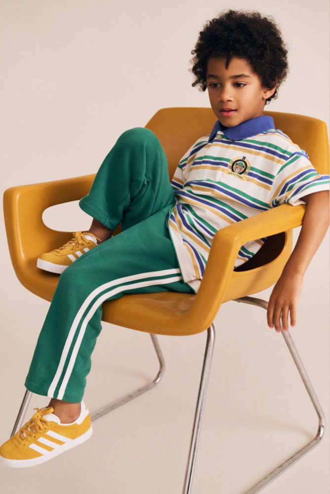 Young boy in a striped polo shirt and green track pants sitting relaxed in a yellow molded chair against a white studio background.