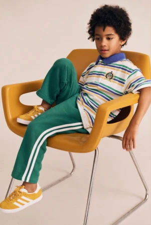 Young boy in a striped polo shirt and green track pants sitting relaxed in a yellow molded chair against a white studio background.